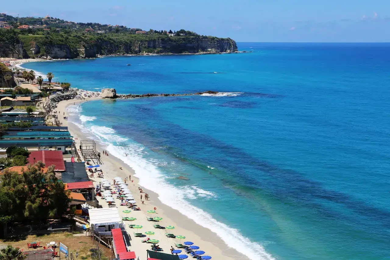 Plage animée à Tropea, Italie, avec des parasols colorés, des baigneurs et des falaises verdoyantes surplombant la mer d'un bleu éclatant.