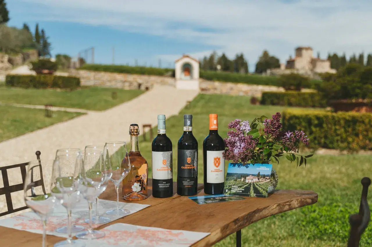Vino e bicchieri sul tavolo di legno, con la campagna toscana e la Tenuta Casenuove sullo sfondo.