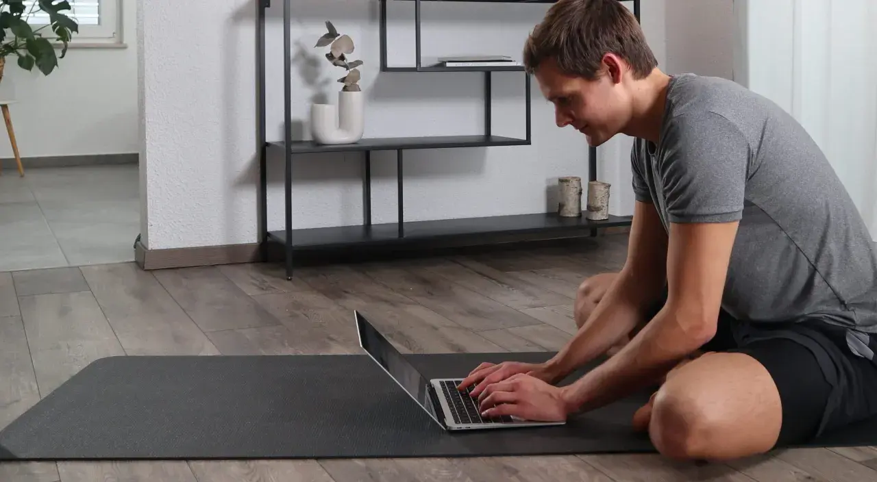 Mann auf Yogamatte tippt auf Laptop, um den Buckel wegtrainieren.