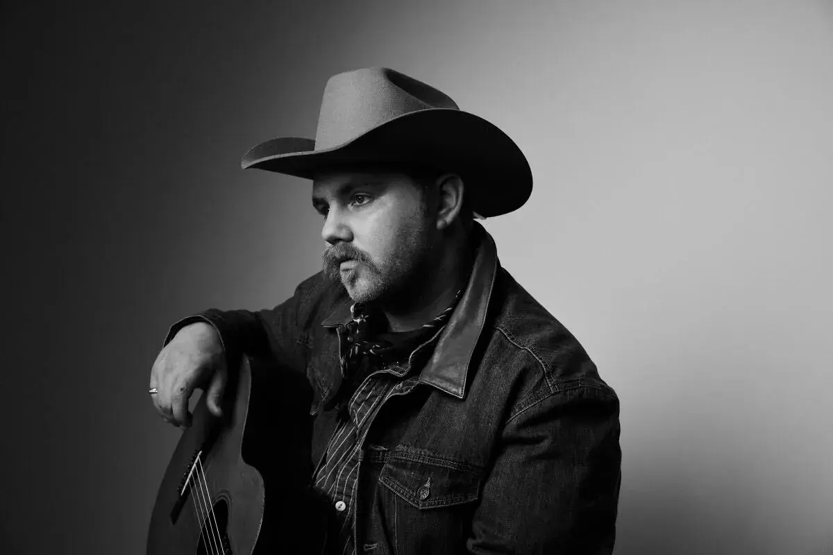 Ernest Keith Smith, a thoughtful country musician, poses with his guitar, wearing a cowboy hat and denim jacket.