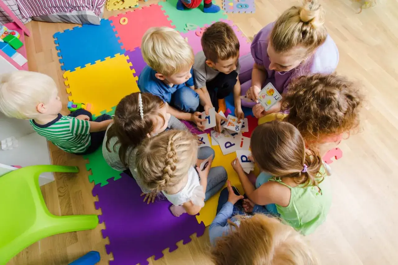 Dzieci i dorosła siedzą na kolorowych puzzlach, grając w karty z literami.