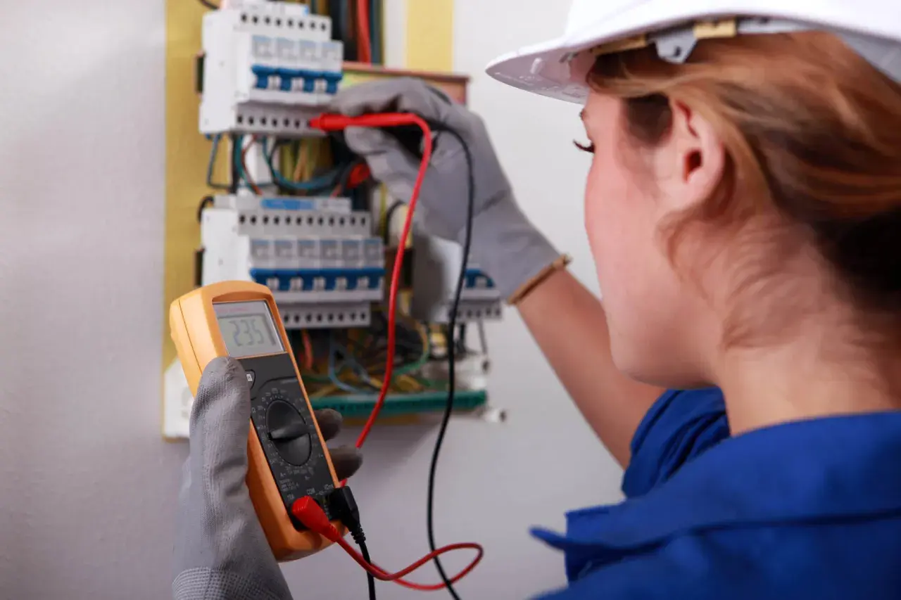 Kobieta w białym kasku wykonuje przegląd instalacji elektrycznej, używając multimetru do pomiaru napięcia.