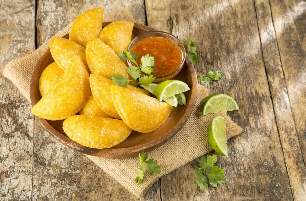 Empanadas colombianas doradas en plato de madera con salsa, limones y cilantro sobre mesa rústica.