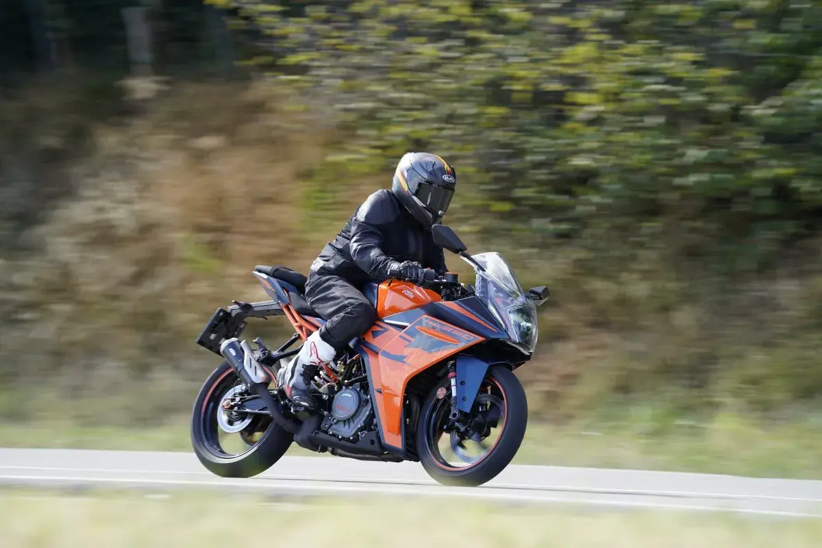 Motociclista con casco y traje negro conduce una KTM RC 390 CV naranja y azul por una carretera con árboles borrosos al fondo.
