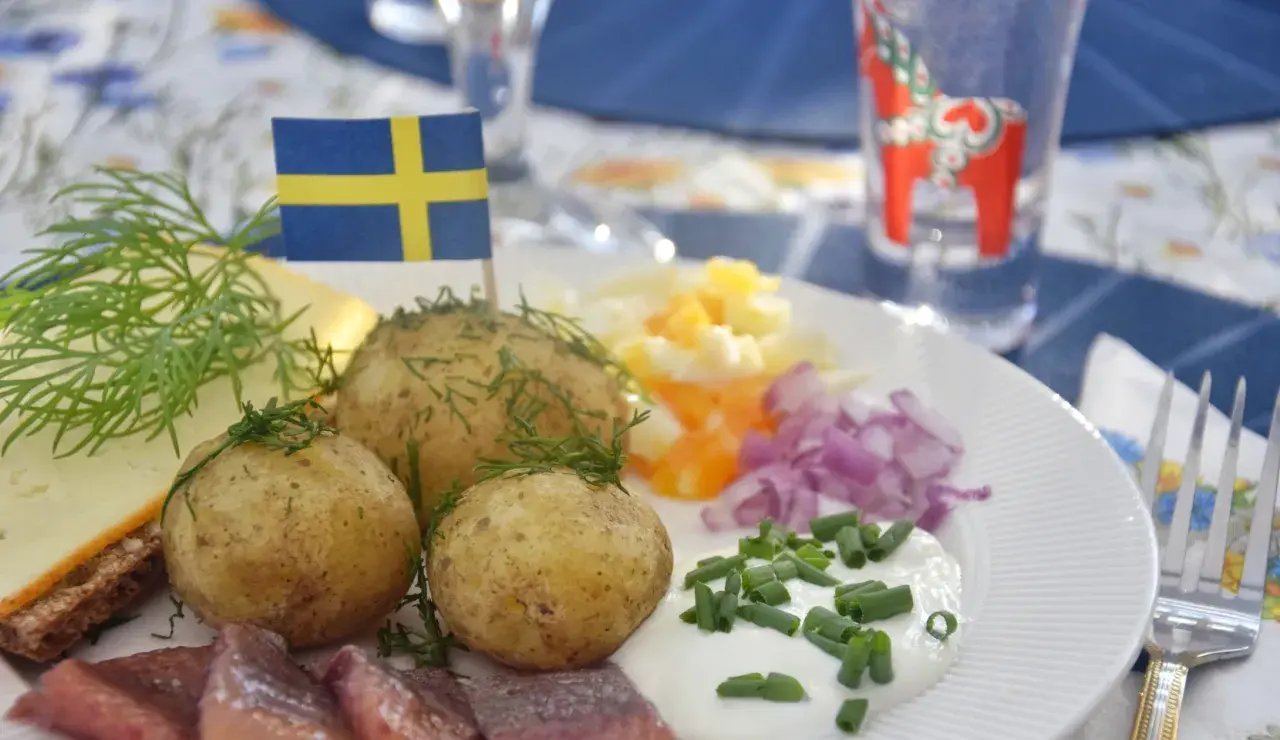 En tallrik med klassisk midsommarmat: sill, färskpotatis, ost, gräddfil och rödlök. En svensk flagga vajar stolt.