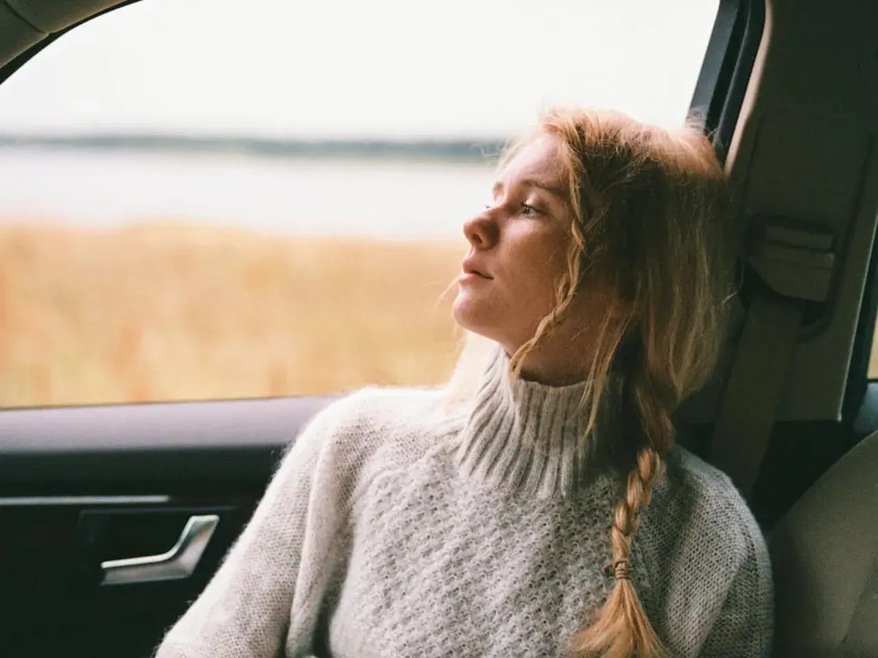 Ragazza con capelli biondi raccolti in acconciature trecce laterali, guarda fuori dal finestrino dell'auto, immersa nei suoi pensieri.
