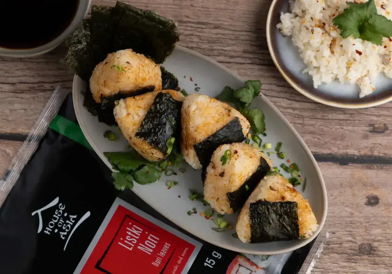Pięć onigiri z ryżem, udekorowanych listkami nori i szczypiorkiem, obok opakowania nori i miseczki z ryżem.
