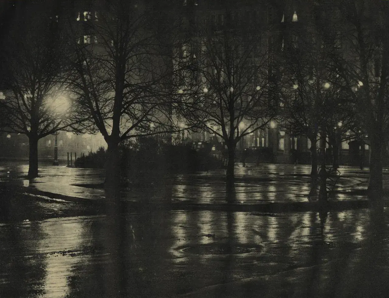 Nocny park, mokre alejki odbijające światła latarni i okien. Piktorializm w fotografii oddaje nastrój i atmosferę.