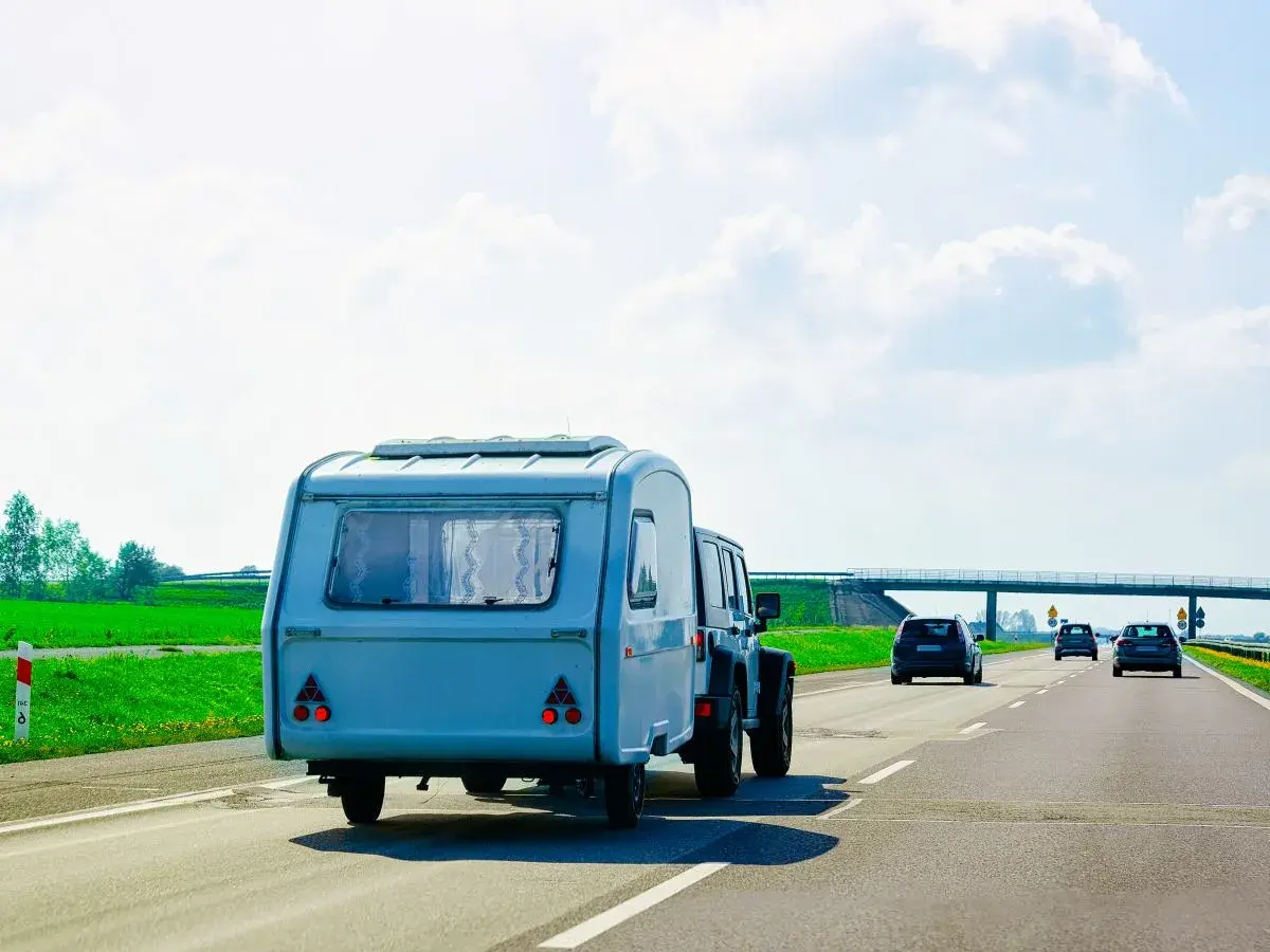 Samochód terenowy ciągnie dmc przyczepy kempingowej po autostradzie. W tle zielone pola i most.