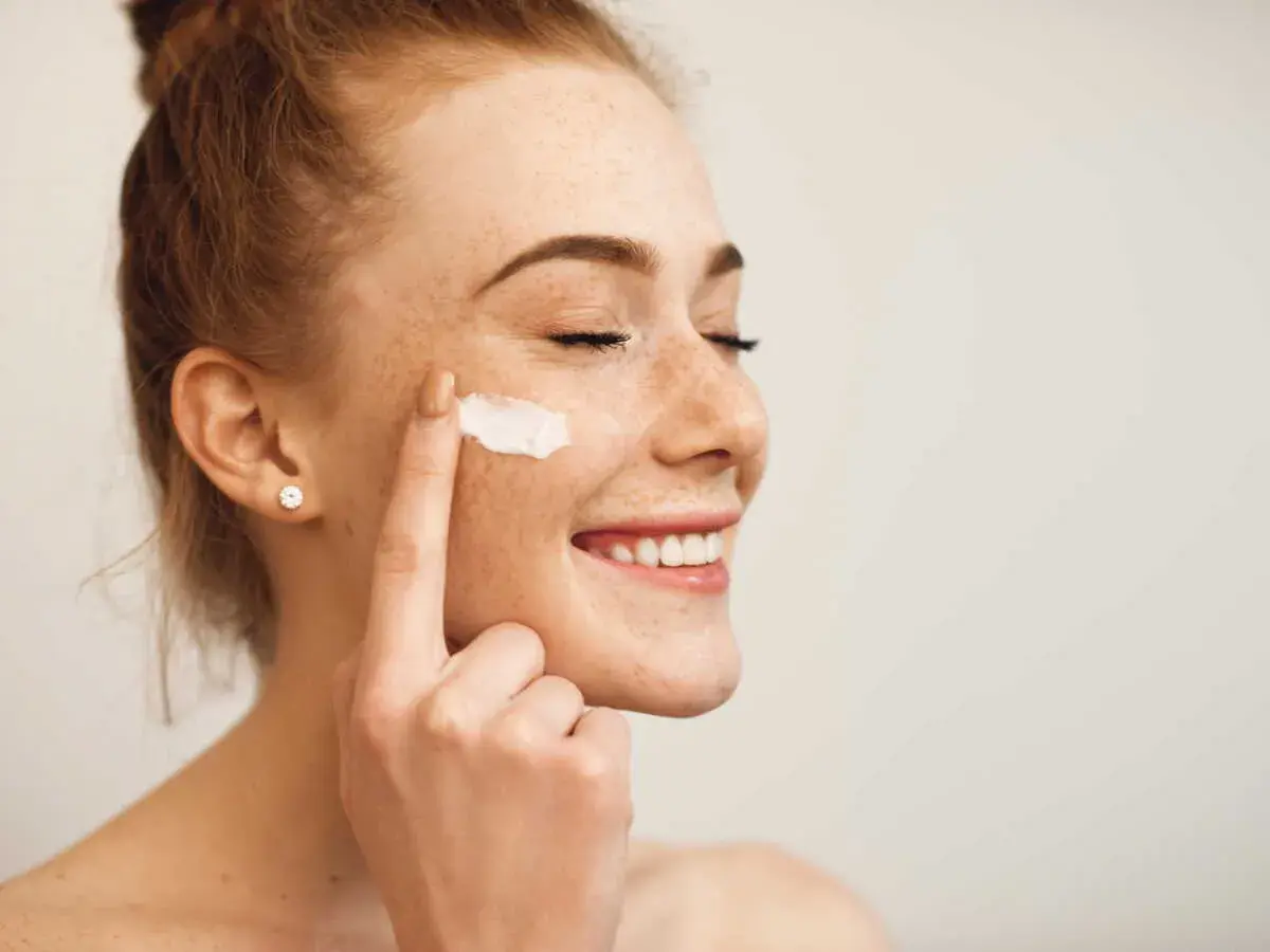 Une jeune femme rousse souriante, les yeux fermés, applique de la crème sur sa joue. Elle montre comment appliquer crème visage pour une peau éclatante.