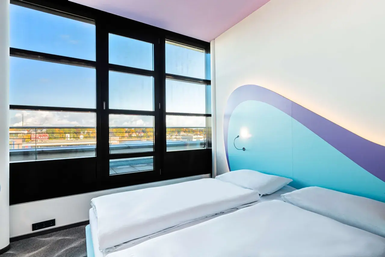 Pokój z dwoma łóżkami w **Radisson Blu Hotel Rostock**. Duże okno z widokiem na miasto i wodę. Nowoczesne, jasne wnętrze z kolorowym zagłówkiem.