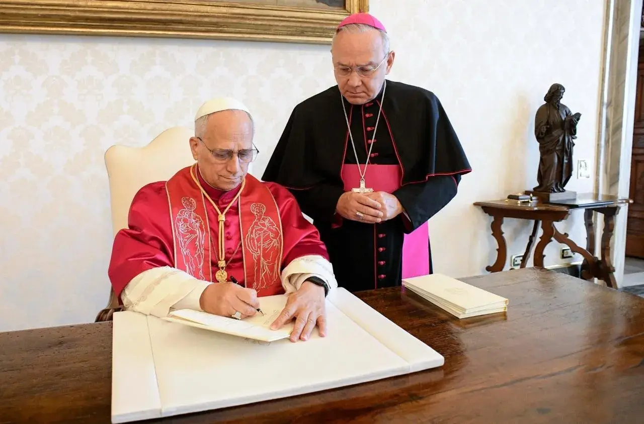 Biskup w purpurowej szacie podpisuje dokument, podczas gdy inny duchowny w czerni i różu stoi obok, obserwując. To ważny moment, być może podpisanie adhortacji.