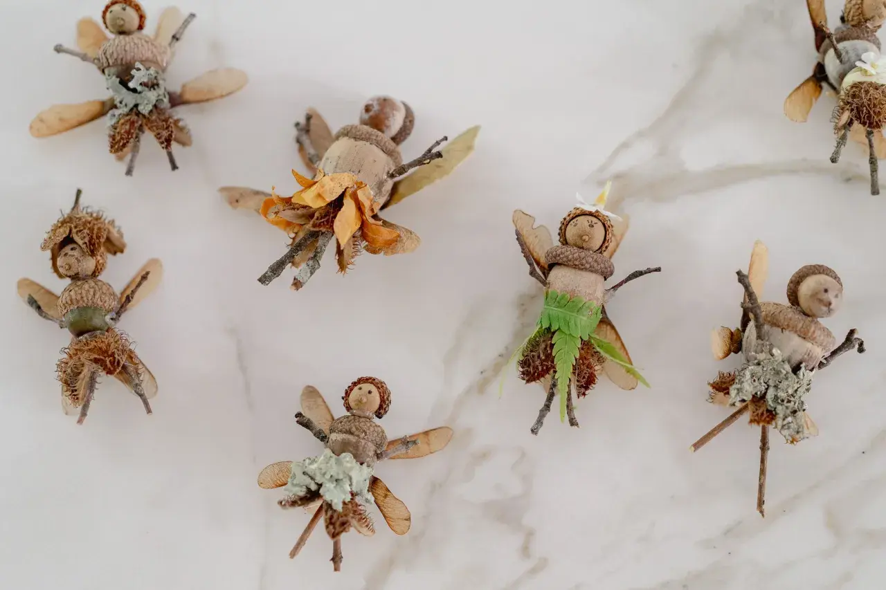 Figuras de hadas hechas con bellotas, ramas y hojas sobre fondo blanco.