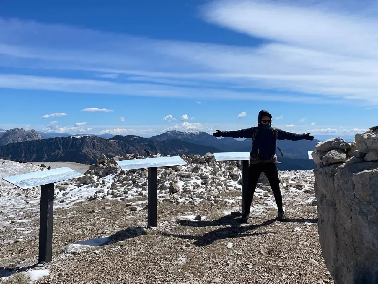 Persona amb els braços oberts al cim del Pedra dels Quatre Batlles, amb paisatge muntanyós i cel blau.