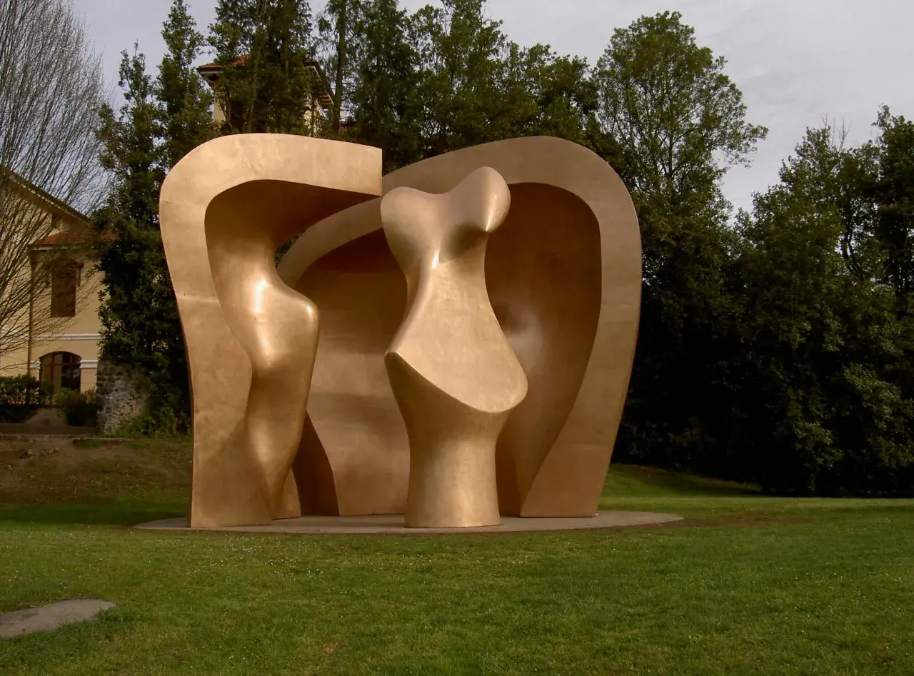 Escultura abstracta dorada en el parque de los pueblos de Europa, con formas curvas y orgánicas, rodeada de césped y árboles frondosos.