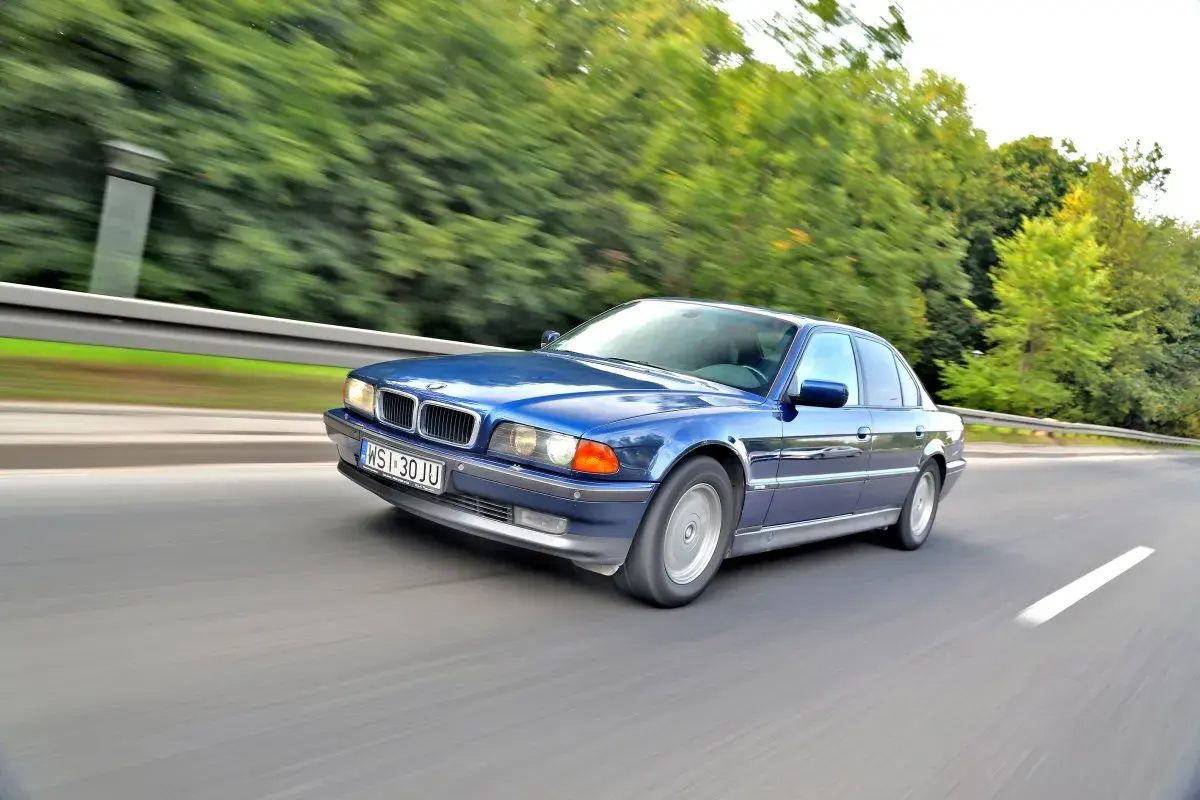 Błękitne BMW 750i pędzi po drodze, otoczone zielenią drzew.
