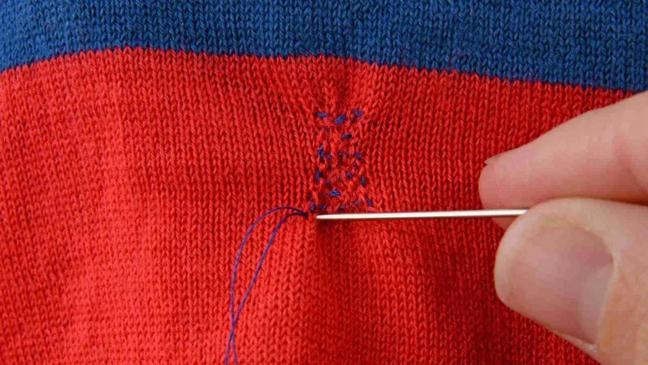 Main d'une personne utilisant une aiguille et du fil bleu pour comment recoudre un tissu déchiré sur un vêtement rayé rouge et bleu.