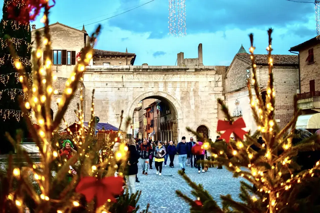 Luci natalizie illuminano un'antica piazza a Senigallia, con persone che passeggiano tra alberi addobbati. Atmosfera magica di Natale.