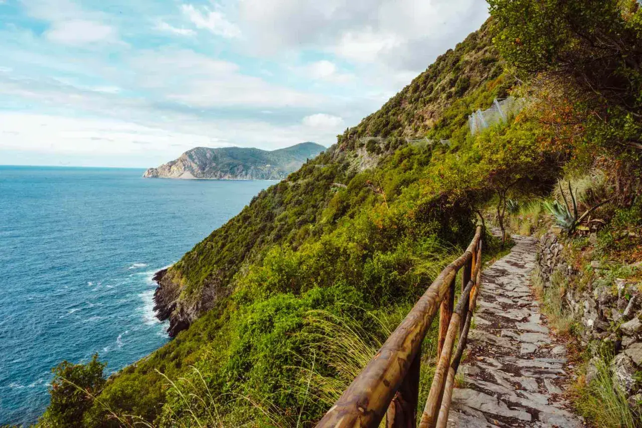 A stone path winds along the coast for Cinque Terre hiking, with lush green hills and the blue sea stretching to the horizon.