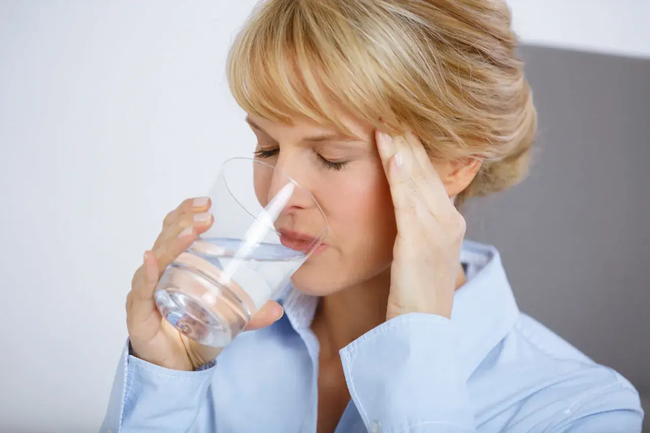 Frau mit Kopfschmerzen trinkt Wasser, hofft auf Linderung durch Magnesium.