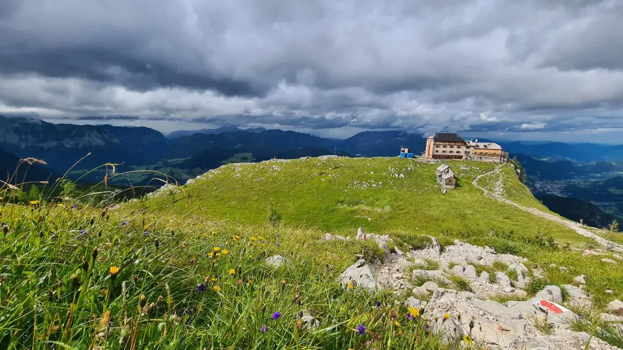 Watzmannhaus Wanderung: Ein Berggasthof thront auf einem grünen Kamm, umgeben von blühenden Almwiesen und dramatischen Wolken.