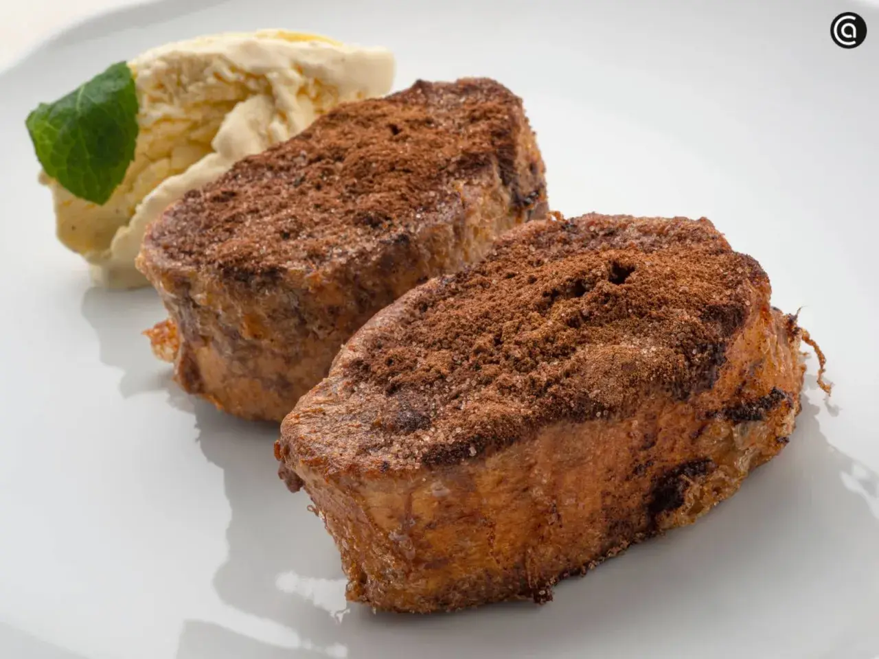 Dos torrijas de chocolate espolvoreadas con cacao y azúcar, acompañadas de helado de vainilla y una hoja de menta.