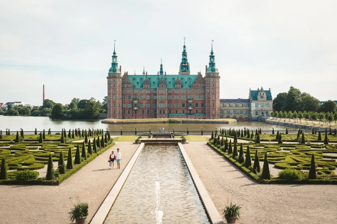 Ett imponerande slott i Danmark, omgivet av vatten och med en vacker barockträdgård. Ett par promenerar längs en damm framför slottet.