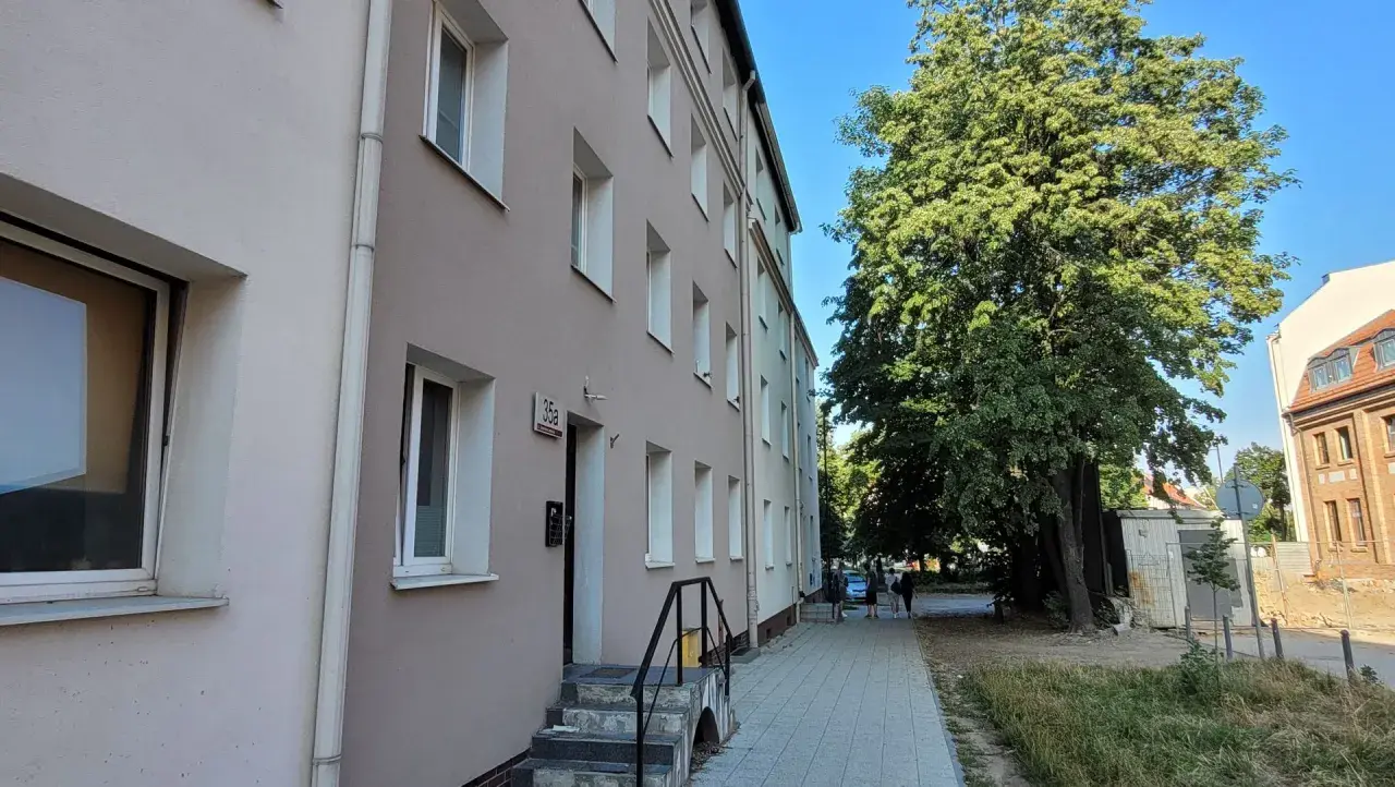 Budynek mieszkalny z oknami i drzwiami, obok drzewa i chodnik. To miejsce może być związane z art. 18 ustawy o ochronie praw lokatorów.