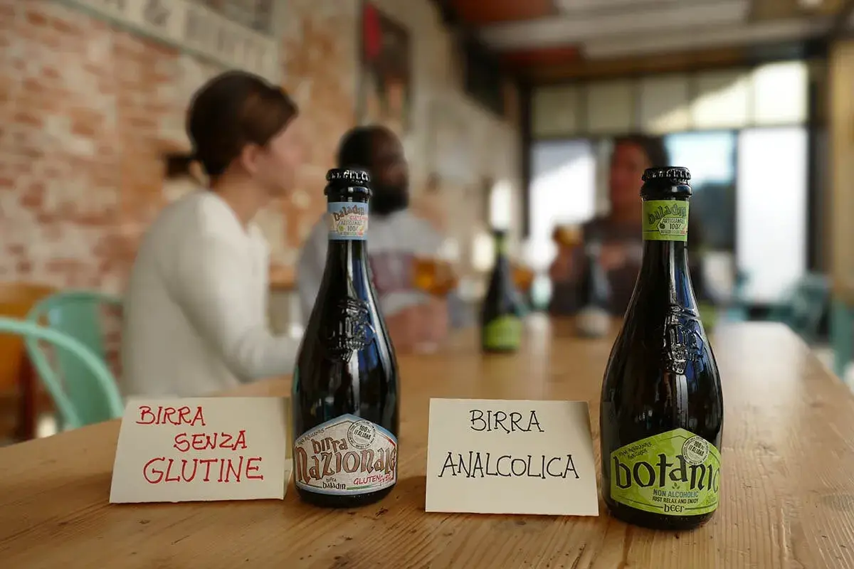 Bottiglie di birra sul tavolo. Una indica "Birra senza glutine", l'altra "Birra analcolica". Attenzione: la birra contiene lattosio.