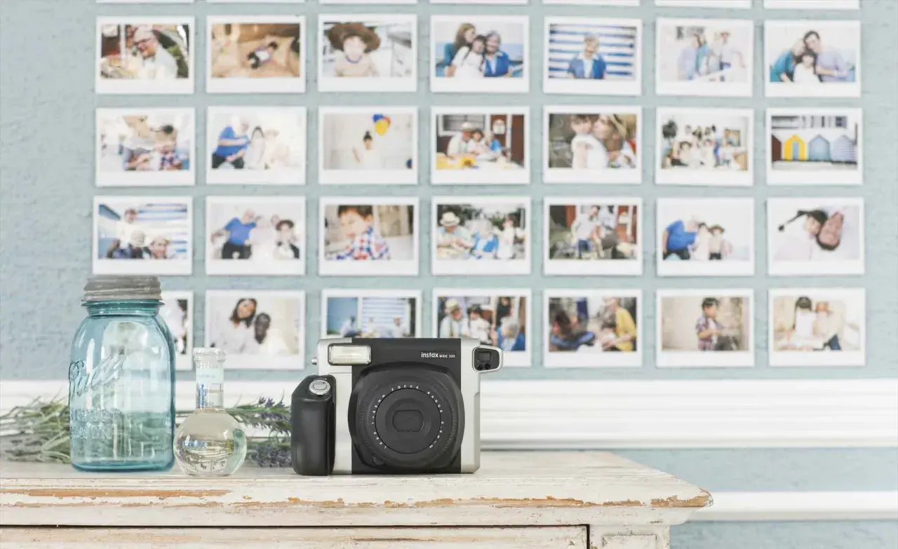 Ściana ozdobiona rzędem zdjęć z polaroida, przedstawiających rodzinne chwile. Obok aparat Instax, słoik i buteleczka.