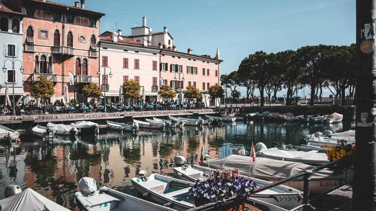 Barche ormeggiate nel porto di Desenzano del Garda, con edifici colorati e alberi sullo sfondo.