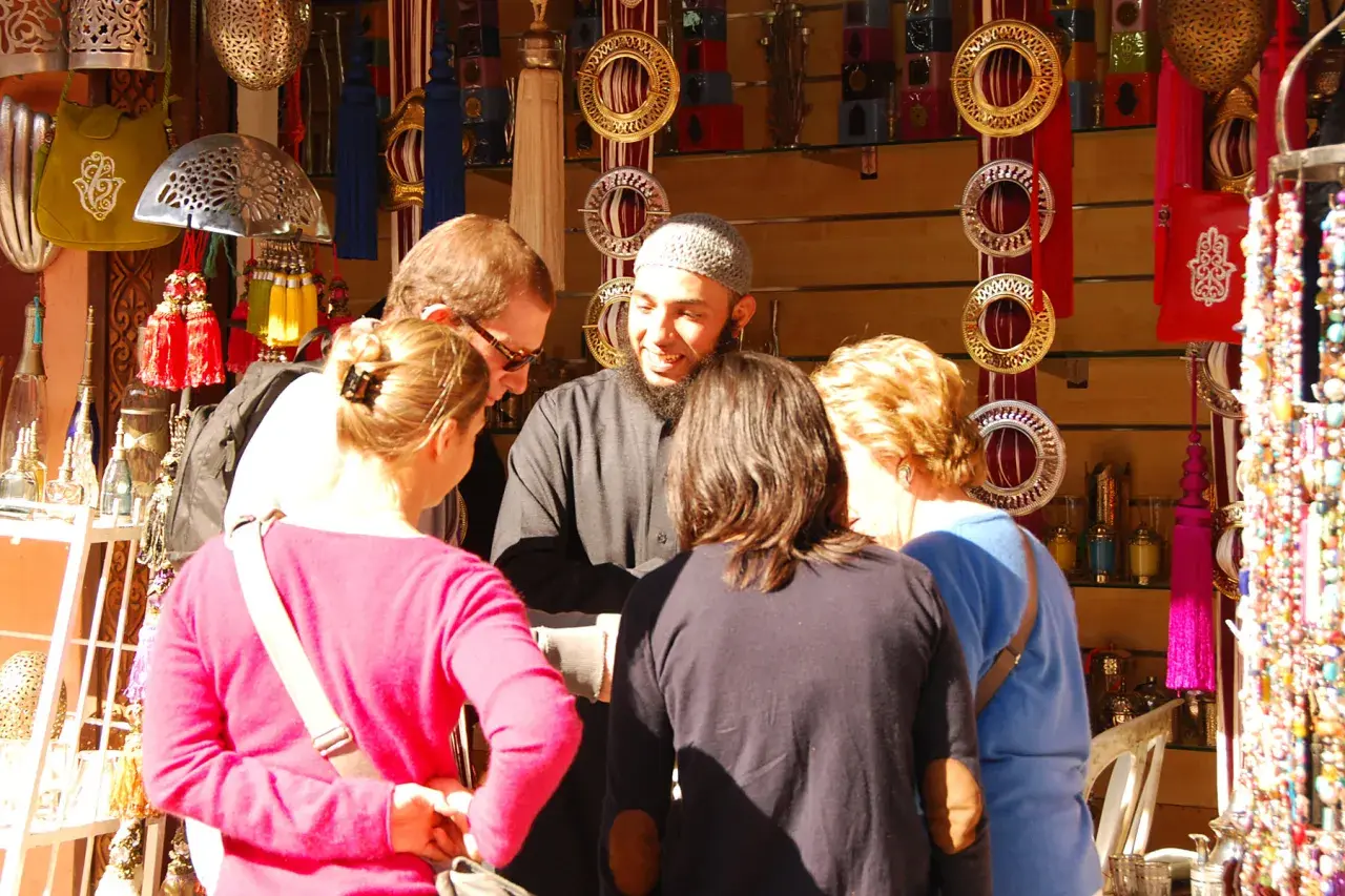 Un groupe de touristes discute avec un vendeur dans un souk de Marrakech, entouré de lanternes, de bijoux et de textiles colorés.