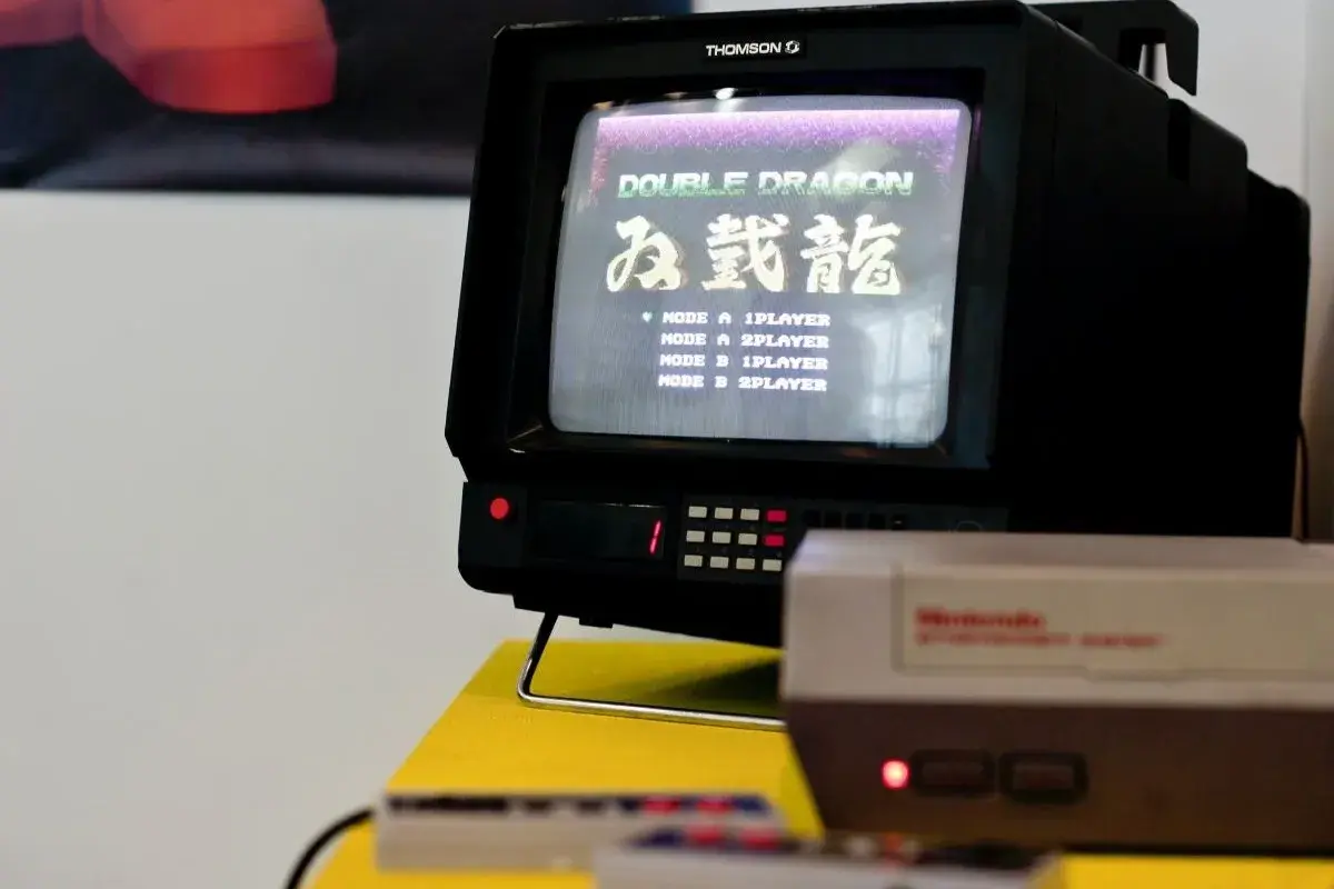 Stary telewizor Thomson z grą Double Dragon. Na ekranie widoczna lista trybów gry, np. "MODE A 1PLAYER". Obok konsola Pegasus.