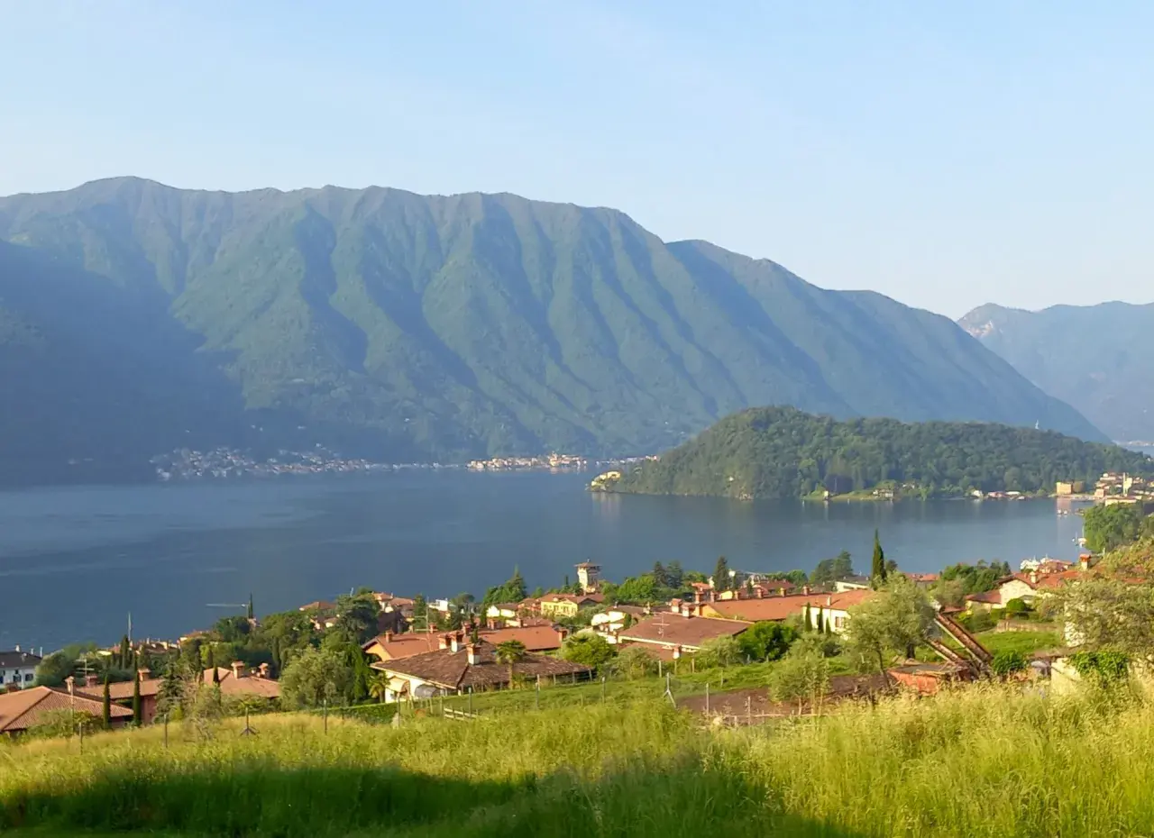 Villaggio sulle rive del lago più grande d'Europa, con montagne verdi sullo sfondo e un prato fiorito in primo piano.