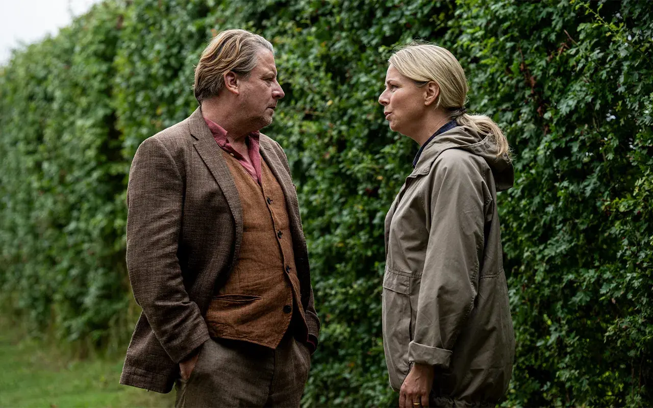 Två personer står och pratar intensivt framför en grön häck. Mannen bär kavaj och väst, kvinnan en jacka. En scen från en dansk kriminalserie på TV4.
