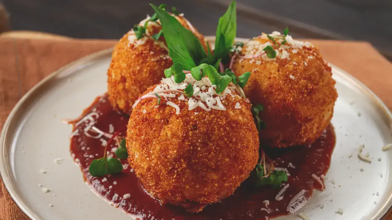 Trois arancini, spécialité italienne, dorés et croustillants, servis sur une sauce rouge avec des herbes fraîches et du parmesan râpé.