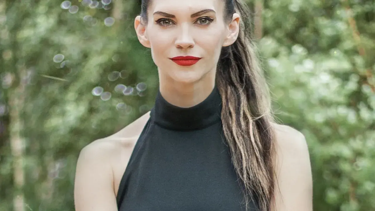 Małgorzata Oliwia Sobczak w czarnej, eleganckiej sukience, z burzą włosów i wyrazistym makijażem, na tle zieleni.
