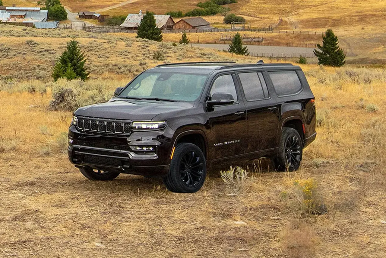 Ciemny Jeep Grand Wagoneer na tle wiejskiego krajobrazu z budynkami i drzewami.