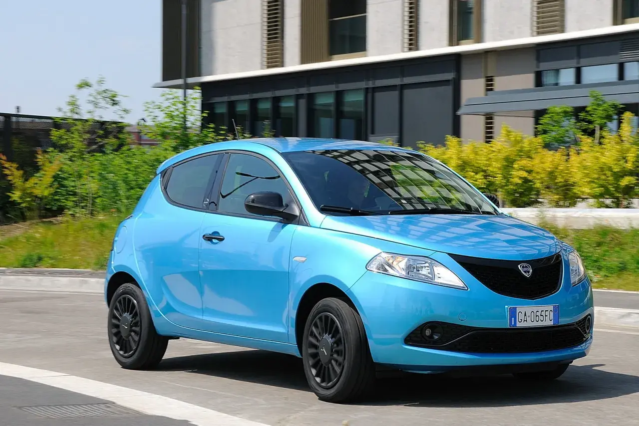Lancia Ypsilon 1.3 Multijet Diesel azzurra in movimento, con sfondo di edifici moderni e vegetazione.