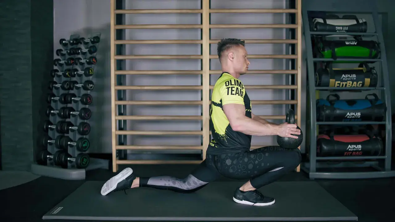 Mężczyzna wykonuje ćwiczenia mobilizacyjne z kettlebell, pogłębiając wykrok. W tle stojaki z hantlami i fitbagami.