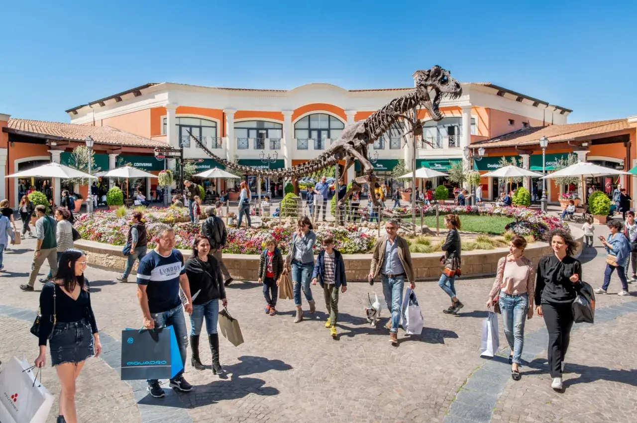 Un T-Rex scheletrico domina il centro del campo rom Castel Romano, circondato da negozi e gente che fa shopping.