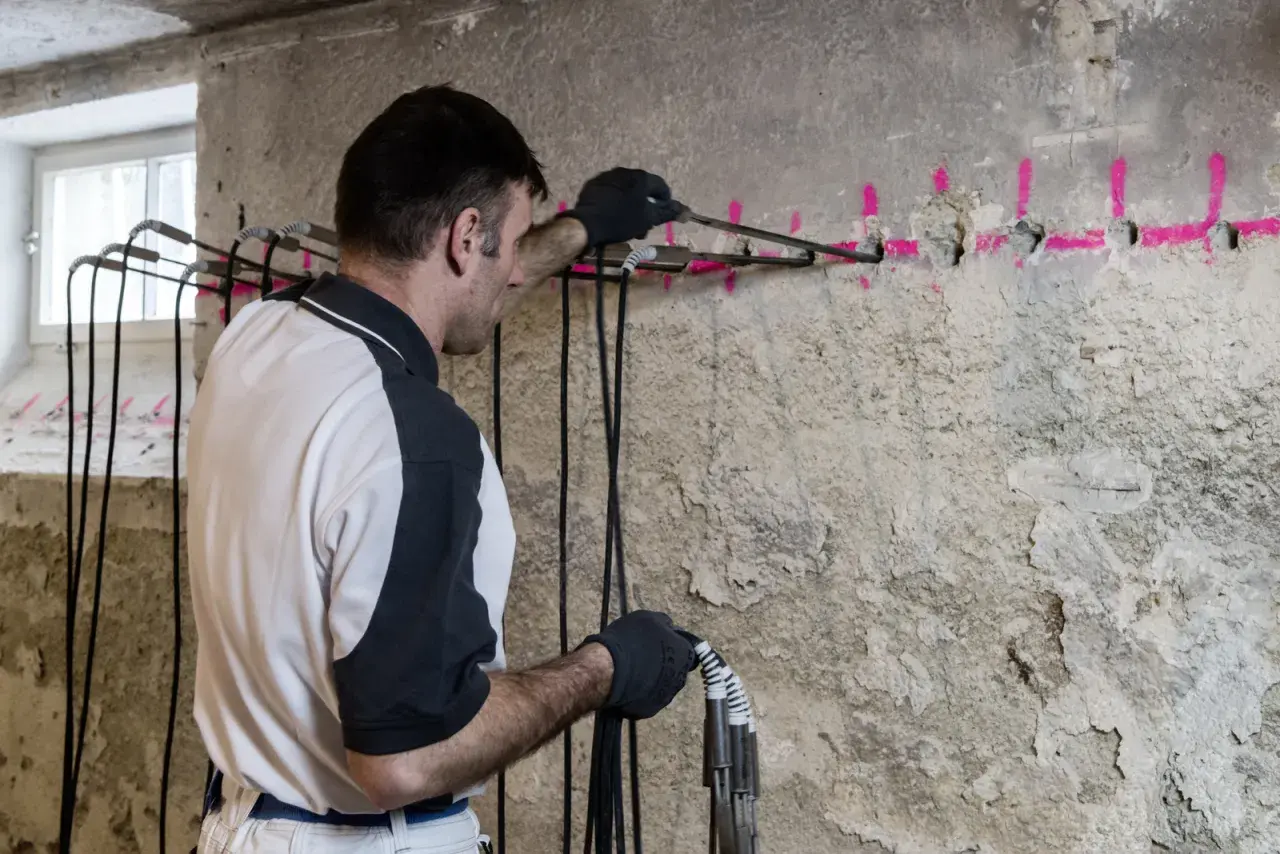 Mann führt eine Horizontalsperre im Mauerwerk durch. Bohrlöcher mit pinker Markierung und Kabel sind sichtbar.