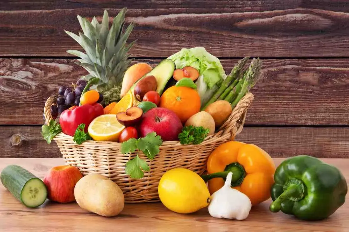 Cesto pieno di frutta e verdura fresca: ananas, uva, mele, arance, limoni, pomodori, peperoni, patate, cetrioli, asparagi e lattuga.
