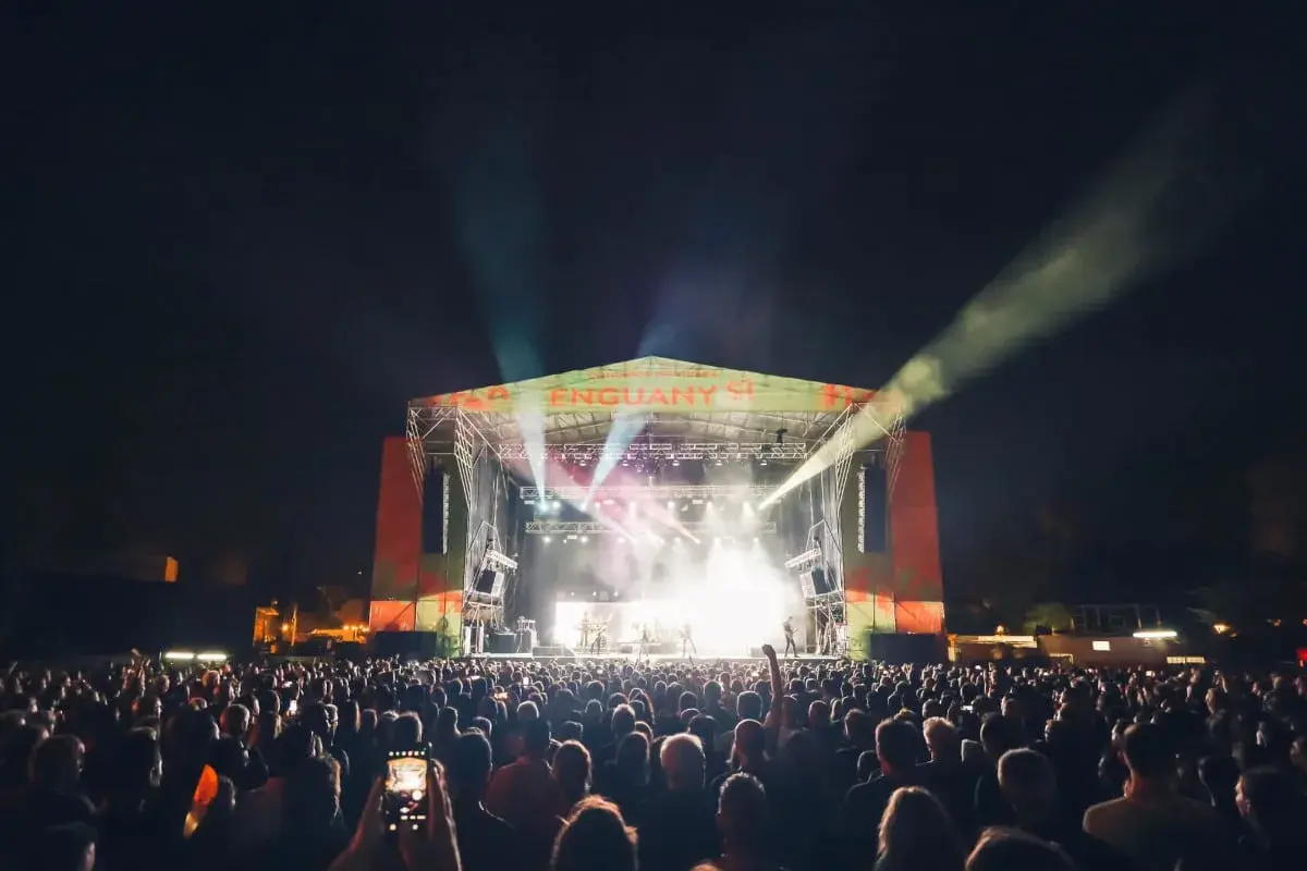 Multitud disfrutando de los **concerts de Vivers** bajo un cielo nocturno iluminado por focos y la energía de la banda en el escenario.
