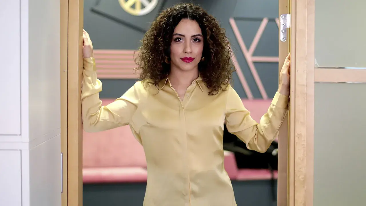 Helya Riazat, med lockigt mörkt hår och rött läppstift, står i en dörröppning i en guldig blus.