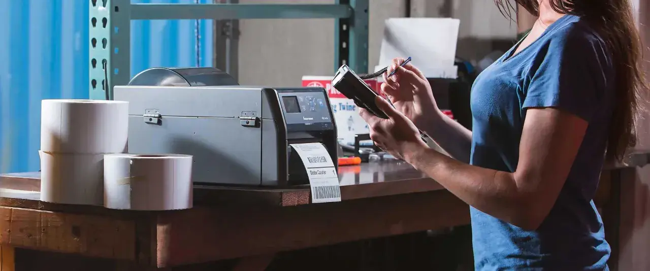 Frau bedient Drucker für Etiketten und Handscanner. Dies ermöglicht effizientes belegloses kommissionieren im Lager.