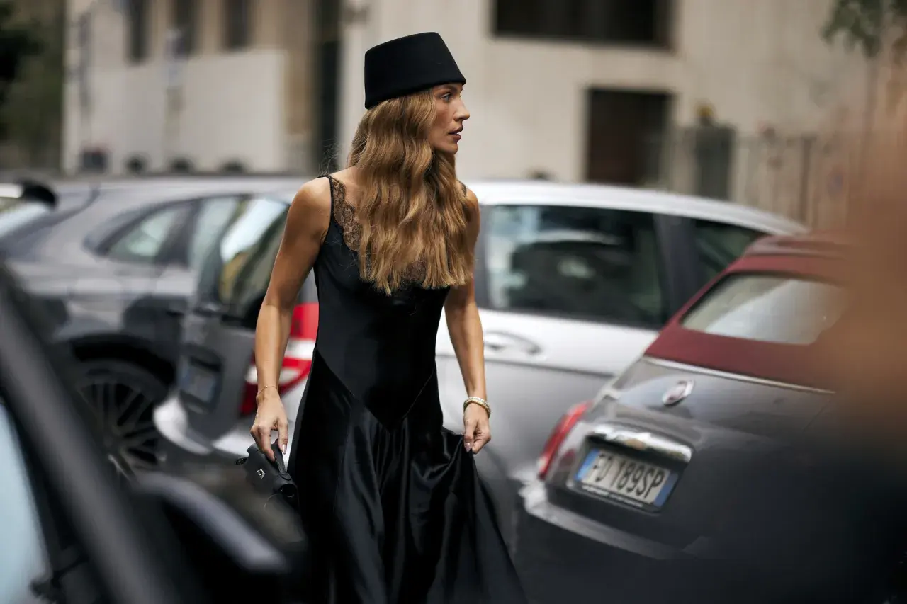 Mujer con sombrero negro y vestido de seda, su pelo en pausa, camina entre coches aparcados.