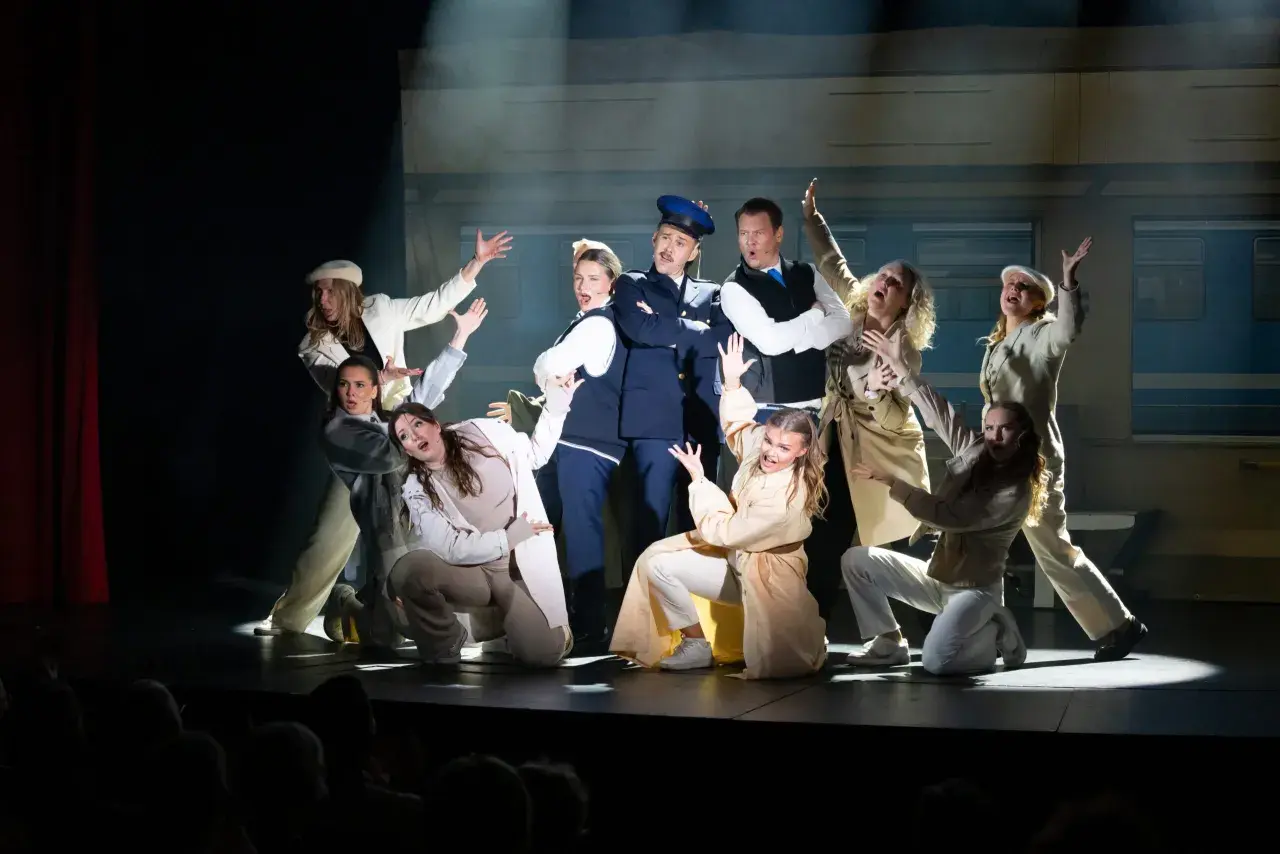 Ensemblen i en svensk revy, med en polisman i centrum, gestikulerar dramatiskt på scenen framför en tågvagnsbakgrund.