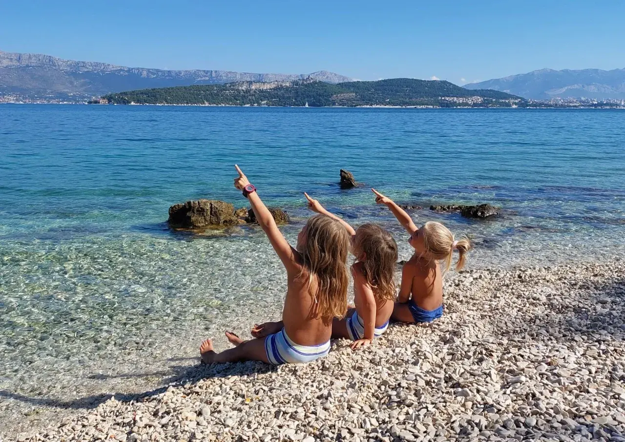 Trzy dziewczynki siedzą na kamienistej plaży w Chorwacji, wskazując palcami na morze. Idealne miejsce, gdzie do Chorwacji z dziećmi.