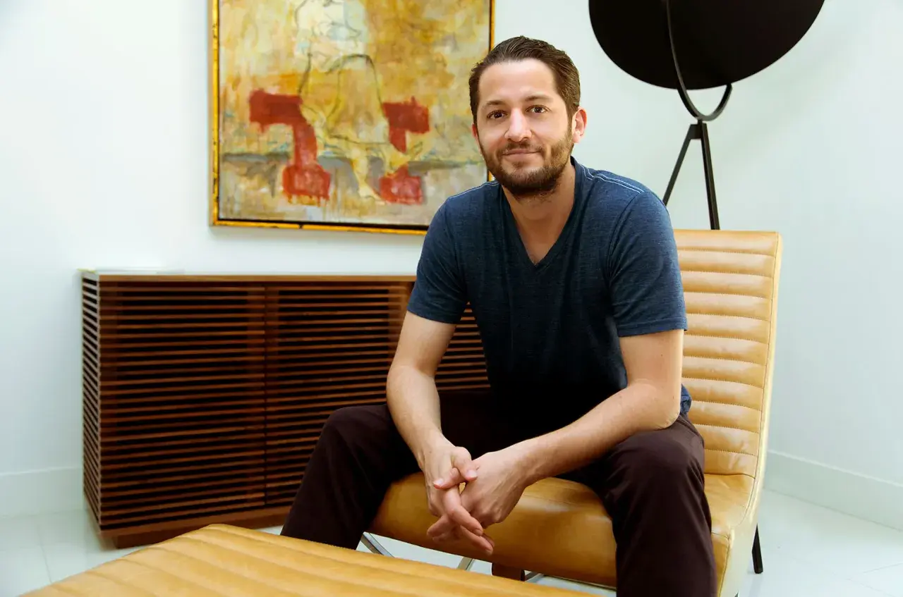 Ian Kirkpatrick, a man with a beard and a blue t-shirt, sits in a tan leather chair in a modern room with abstract art.